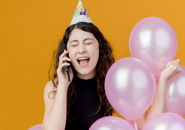 女人年轻漂亮的女人 卷发 戴着节日帽 手持气球 讲着手机 站在橙色的墙上 快乐而兴奋的生日派对抱着说话兴奋
