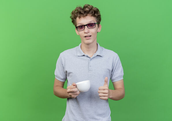 杯子戴着黑眼镜的年轻人穿着灰色马球衫 手里拿着咖啡杯 微笑着竖起大拇指站在绿色的墙上眼镜男人拇指
