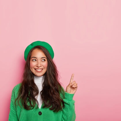 表情帅气的亚洲女士戴着绿色贝雷帽和针织套头衫 手指上方 表情开朗日本人毛衣指向