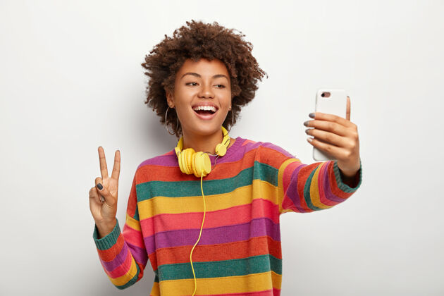 笑无忧无虑的卷发女青年在手机上自拍写真 展现和平姿态 穿着条纹彩色套头衫人肩膀民族