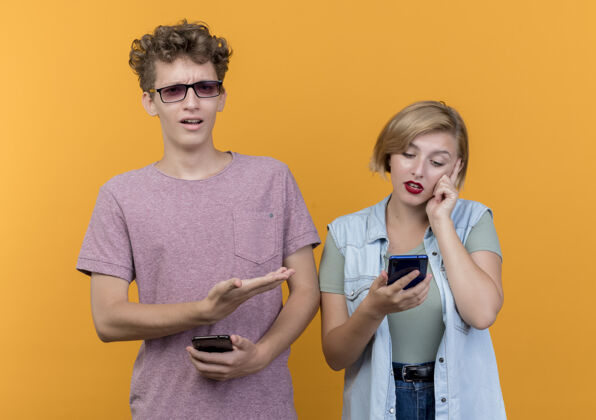 移动年轻漂亮的一对男女穿着休闲服 手持智能手机站在橙色的墙上吵架随便情侣年轻