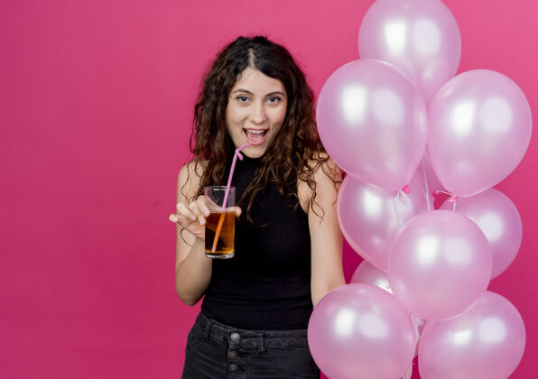 空气一个年轻漂亮的女人 卷发 手里拿着一束气球 手里拿着鸡尾酒 站在粉红色的墙上 开心地笑着抱着鸡尾酒卷发