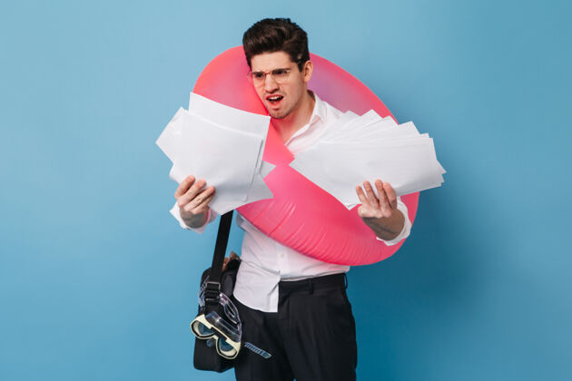 男性不高兴的家伙穿着白衬衫 手里拿着一堆办公用纸深色的男性拿着粉色的橡胶圈在蓝色的空间里摆姿势文件老板优雅