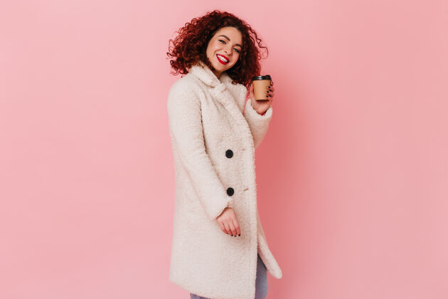 漂亮可爱的黑发女人 穿着浅羊毛外套 带着雪白的微笑 在粉色的空间里捧着一杯硬纸板咖啡卷曲红色嘴唇女性