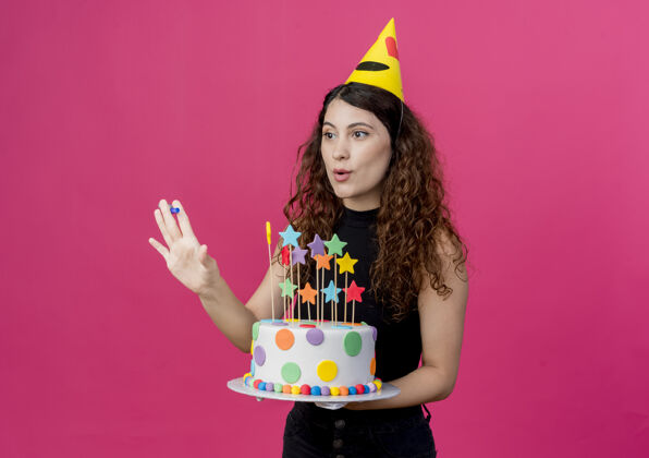 卷发年轻漂亮的女人 卷发 戴着节日帽 手里拿着生日蛋糕 站在粉色的墙上 快乐而积极的生日派对理念帽子年轻女人