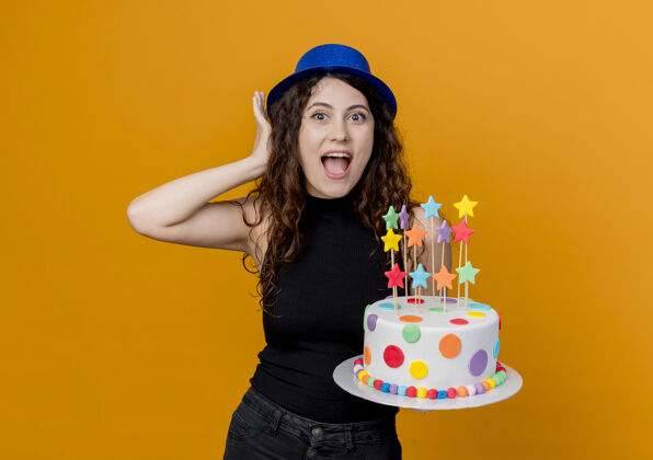 年轻一个年轻漂亮的女人 卷发 戴着节日帽 手里拿着生日蛋糕 站在橘色的墙上 又高兴又惊讶女人惊喜抱着