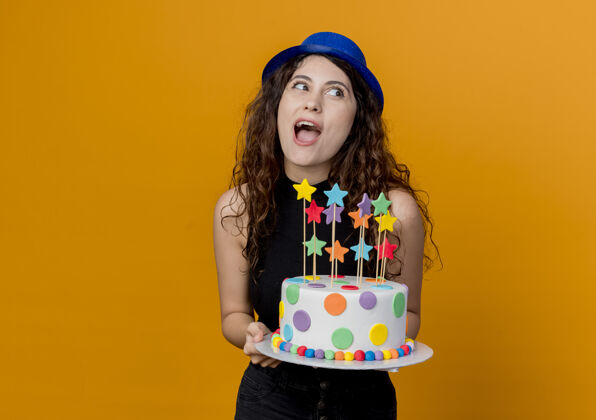 女人一个年轻漂亮的女人 卷发 戴着节日帽 手里拿着生日蛋糕 站在橘色的墙上 又高兴又惊讶蛋糕帽子抱着