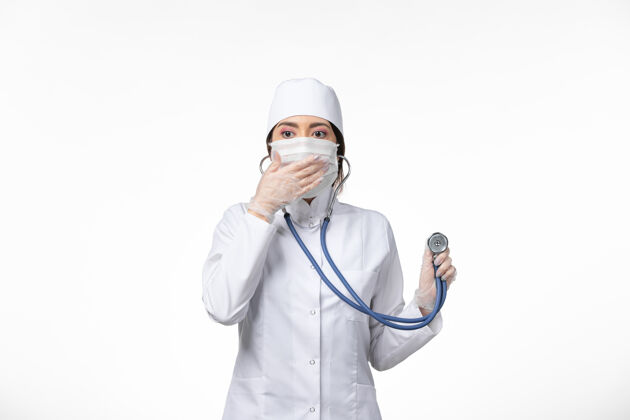 套装正面图女医生身穿白色无菌医疗服 戴口罩 因冠状病毒-使用听诊器对轻白墙病病毒冠状病毒-大流行性疾病病毒医疗大流行