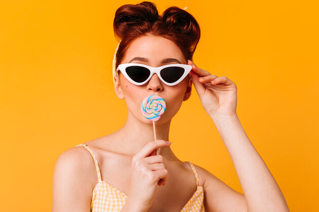 时尚戴着太阳镜舔着硬糖的好女人黄色空间里孤立着一个戴着棒棒糖的小女孩的前视图糖果甜蜜发型