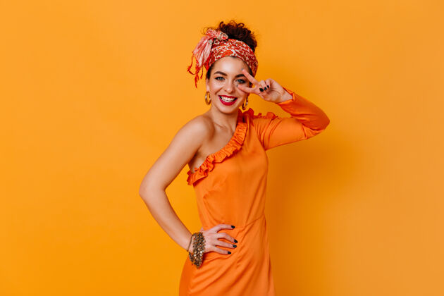 时尚正派女士身着橙色优雅的礼服 头戴时尚的头巾 真诚地微笑着 在橙色的空间里展示着和平的标志头发时尚浪漫