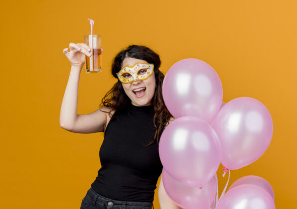 欢呼一位年轻漂亮的女士 卷发 手持一束气球和鸡尾酒 戴着派对面具 站在橙色的墙上 快乐快乐的生日派对理念卷发女人气球