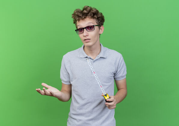 戴着一个戴着黑眼镜 穿着灰色马球衫 拿着卷尺的年轻人站在绿色的墙上做着困惑的手势马球手测量
