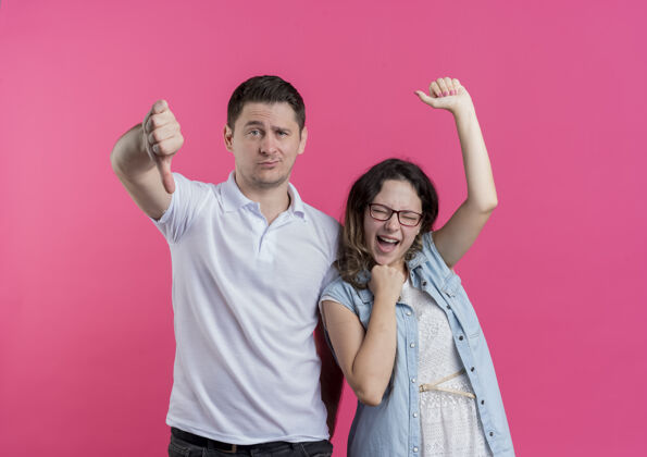 情侣一对年轻夫妇穿着休闲服的男人竖起大拇指 他的女朋友站在粉红色的墙上欢呼雀跃而女朋友男人