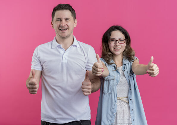 年轻人一对穿着休闲服的年轻夫妇站在粉红色的墙上 兴高采烈地笑着竖起大拇指表情休闲表演