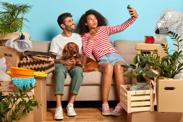 运动鞋家庭情侣自拍的室内镜头 非洲女人在智能手机的摄像头里吹气亲吻 和宠物在舒适的沙发上摆姿势 搬进新的现代公寓 打开箱子房间休闲狗