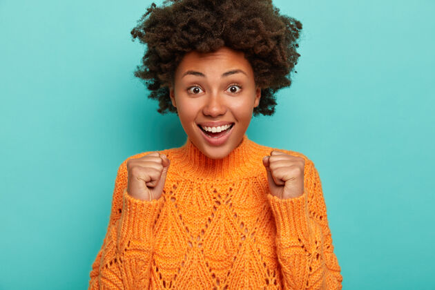 朋友魅力可爱的非洲裔美国女士欢呼胜利 举起紧握的拳头 庆祝某事 笑容灿烂 露出洁白的牙齿 穿着针织橘色毛衣情感非洲女人