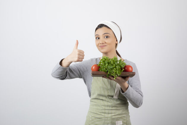 女人微笑可爱的女士模型与新鲜蔬菜木板显示拇指向上拿着围裙女人
