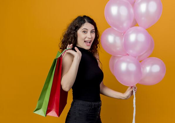 包年轻漂亮的女人 卷发 手持一束气球和纸袋 站在橘色的墙上 微笑着庆祝生日女人微笑卷发
