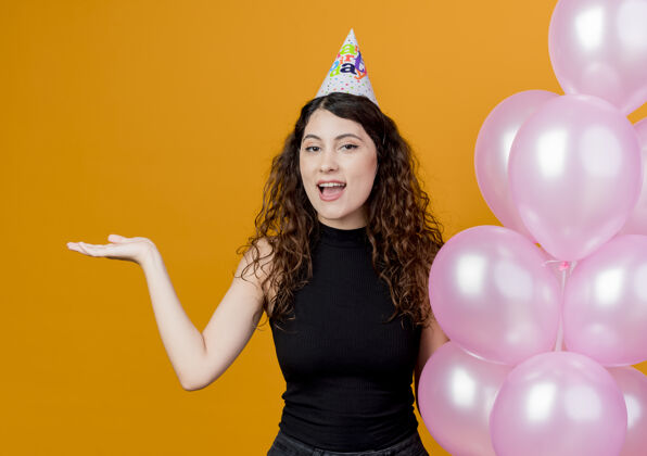 微笑一位年轻漂亮的女士 卷发 戴着节日帽 手持气球 手臂微笑着 站在橙色的墙上 展示生日派对的概念东西站着年轻