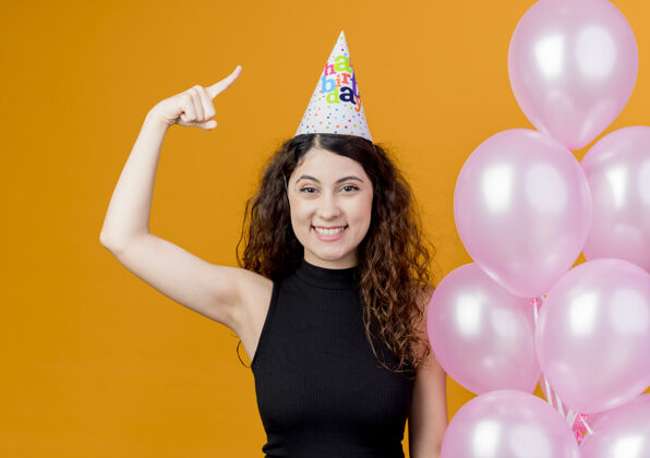 卷发一个年轻漂亮的女人 卷发 戴着节日帽 手里拿着气球 露出食指 快乐而积极的微笑 站在橙色的墙上 欢快地庆祝生日派对目录积极空气