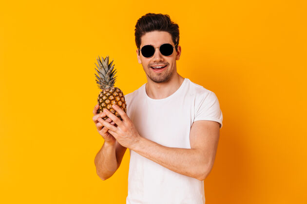 强壮身着白色t恤 戴着太阳镜的男子在橙色空间举着菠萝的肖像人类站立T恤