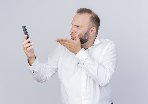 亲吻留着胡子的男人穿着白衬衫拿着智能手机看着屏幕有视频电话吹着吻站在白墙上吹通话穿