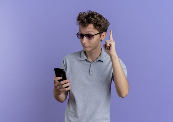 显示穿着灰色马球衫的年轻人看着他的智能手机屏幕 惊讶而高兴地把食指伸到蓝色的墙上 有了新的想法手机男人惊喜