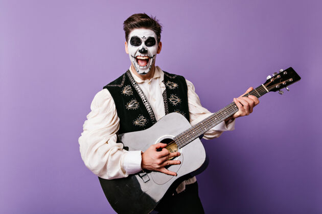 骷髅兴奋的黑发男人带着僵尸妆在紫色的墙上唱歌穆尔特家伙弹吉他大笑的室内镜头娱乐情感墨西哥