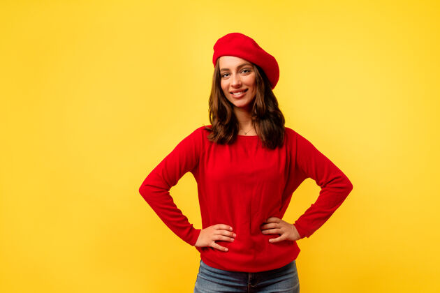 姿势身着红色时尚套头衫和贝雷帽的年轻欧洲迷人女士在黄色墙壁上摆姿势的内部照片笑快乐年轻