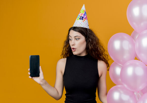 帽子一个年轻漂亮的女人 卷发 戴着节日帽 手里拿着气球 展示着智能手机 看着它 站在橙色的墙上 困惑着生日派对的概念空气抱着看