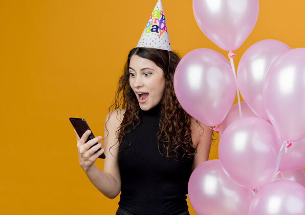 卷曲一位年轻漂亮的女士 头上戴着节日帽 手里拿着气球 看着智能手机的屏幕 站在橘色的墙上 快乐而兴奋的生日派对概念女人帽子站着