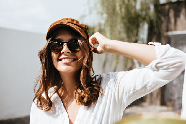 现代一个穿着白色浅v领上衣的快乐女人微笑着看着镜头戴着帽子和眼镜的女孩对着建筑和树木摆姿势姿势裤子白色建筑