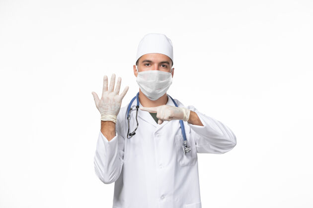 病毒正面图：男医生穿着医疗服 戴着口罩 用听诊器对着白色办公桌上的柯维病大流行病毒制服专业听诊器