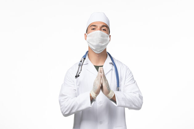 口罩正面图：男医生穿着医疗服 戴着防冠状病毒口罩 用蓝色听诊器对着白墙冠状病毒大流行病毒祈祷病毒男性大流行