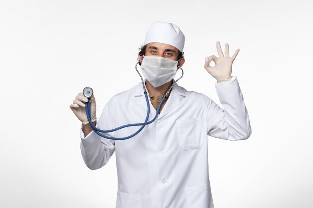 医疗正面图：男医生穿着医疗服 戴着口罩作为防护罩 用听诊器在白色墙壁上观察疾病-病毒-大流行大流行外套西装