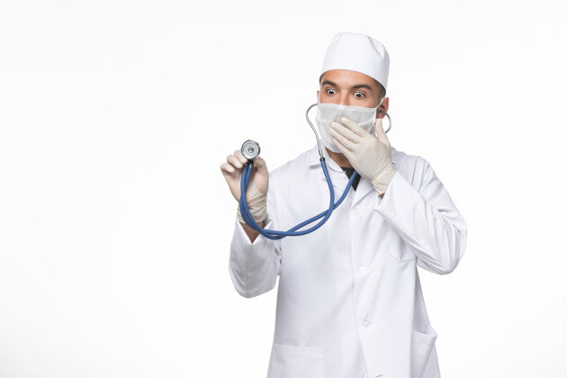 视图正面图：男医生穿着医疗服 戴着口罩 在白色办公桌上用听诊器抵御冠状病毒-冠状病毒疾病大流行病毒男性听诊器