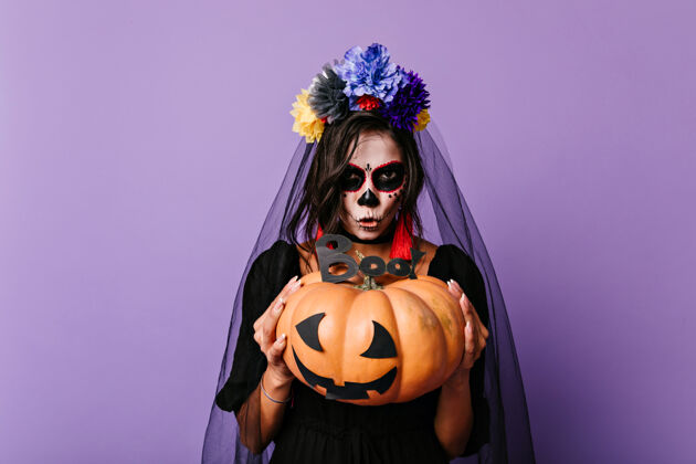 化妆吓人的死新娘抱着南瓜万圣节时 一个戴着黑色面纱的欧洲女人在紫色的墙上摆姿势墨西哥头骨万圣节