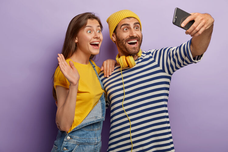 女人喜出望外的女朋友和男朋友的照片在智能手机上自拍 打视频电话 对着相机挥手 开心的表情 一起玩 在紫色背景下在室内摆姿势肖像紫色情侣