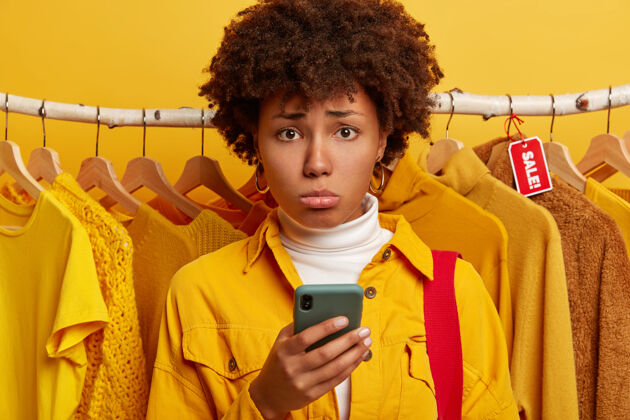 优惠沮丧的非洲女人用智能手机上网购物 不开心 站在衣架上的黄色衣服上网页不满移动