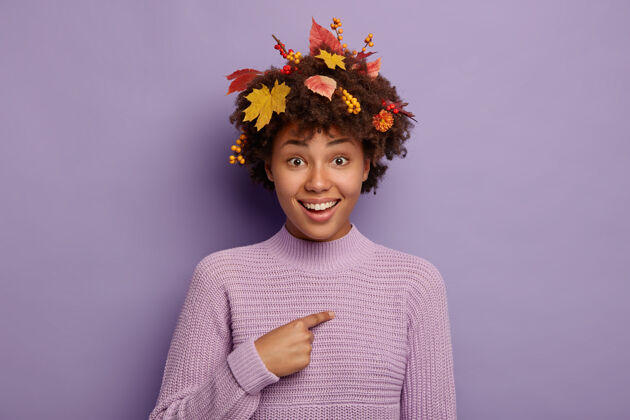 秋天美丽的秋姑娘指着自己 高兴地被接过来参加季节性的节日 穿着暖和的针织毛衣 五颜六色的叶子 浆果和花在头发上 孤立在紫色的背景上站着针织高兴
