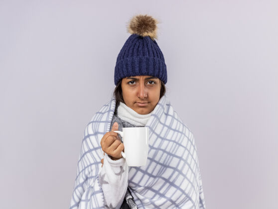 茶未出嫁的生病的年轻女孩戴着冬天的帽子 围巾裹着格子呢 手里拿着一杯隔离在白色上的茶抱着白帽子