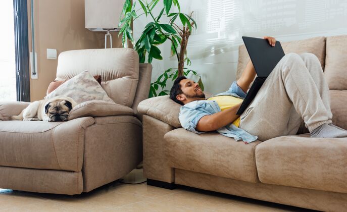 休闲一个男人躺在家里的沙发上用黑色笔记本电脑工作的水平镜头狗沙发人