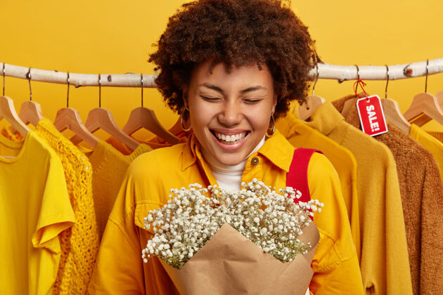 销售照片中的快乐女人手持花束 穿着时髦的黄色夹克 笑容灿烂 欢欣鼓舞 站在衣架上的衣服旁边牙齿微笑单色