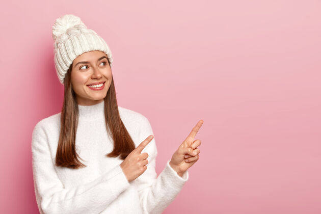 兴奋高兴的黑发女性站在一旁 指着空白的复印空间 穿着冬装 开心地笑着 孤立在粉红色的背景上指点喜悦休闲