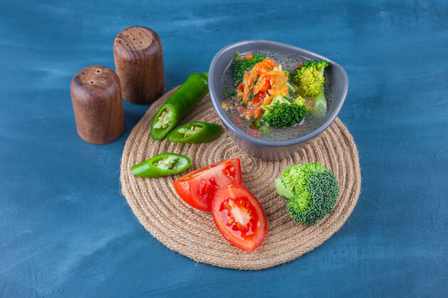 美味碗里的鸡汤和蓝色表面的三脚架上的蔬菜片三叉戟蔬菜碗