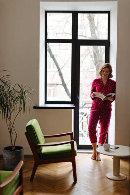 漂亮微笑的欧洲女人在早晨阅读杂志的全景穿着睡衣站在窗边的可爱赤脚女人的室内镜头杯子休闲茶