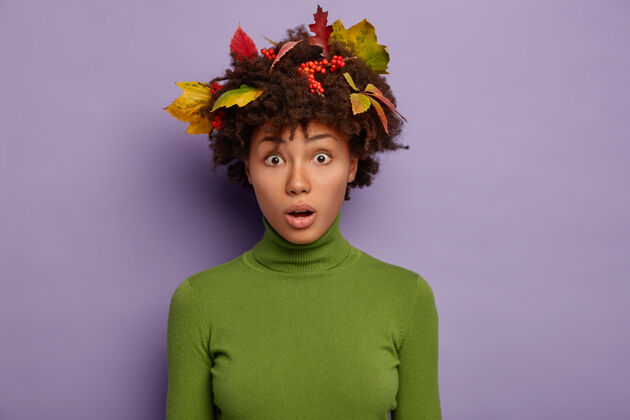 紫罗兰色害怕的非洲裔美国女模特头发上有五颜六色的叶子 穿着绿色的马球衫 隔离在紫色的墙壁上 保持皮肤健康肖像害怕尴尬