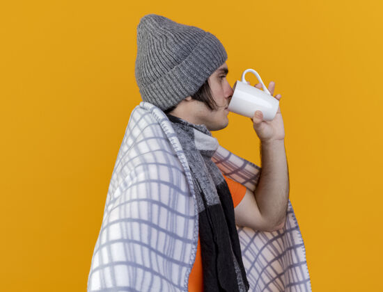 侧写站在侧视图上的年轻病夫戴着冬天的帽子 围巾裹着格子布喝茶格子茶男人