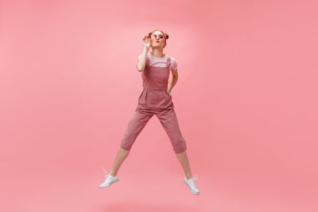 眼睛穿着工作服和白色t恤的快乐女人戴上太阳镜 在粉色背景上跳跃性感卷发头发