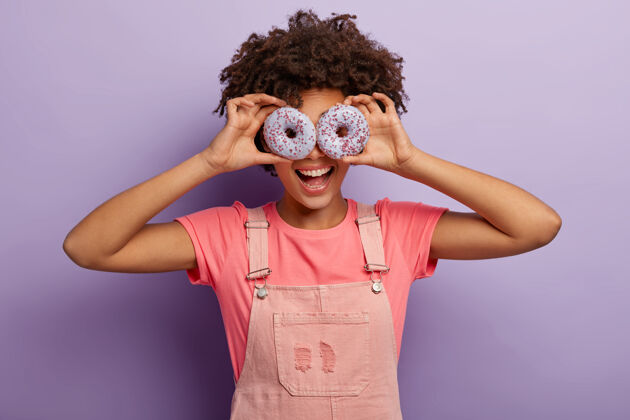 工作服美丽有趣的非裔美国女人眼睛上戴着甜美的紫色甜甜圈 在室内享受美味的甜点 穿着粉色衣服 在紫色背景下与世隔绝节食 垃圾食品 减肥概念室内高兴不健康